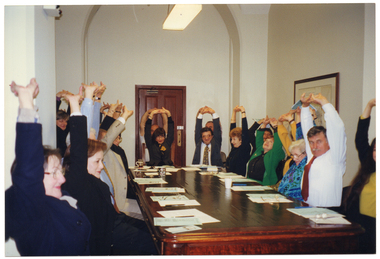 COL photo of a group of people, seated around a long wooden table. They are stretching their arms above their heads with their fingers interlocked and palms turned upwards. Each person has some paperwork on the table in front of them. There are also some pens, a few mugs, a few pairs of spectacles, and a polystyrene cup on the table.