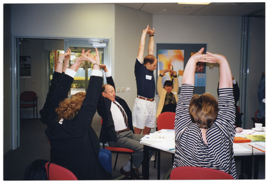 COL photo of a small group of people in a large room. One man is standing, while the others are sitting around some tables. They are all stretching their arms above their heads, with their fingers interlocked and palms turned upwards. In the background, there are some posters on a display board on the wall and a set of glass double doors leading to another room.