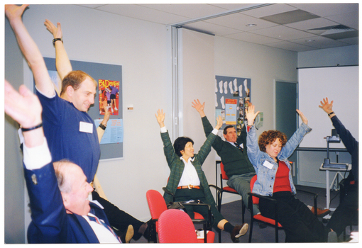 COL photo of a small group of people in a large room. One man is standing, while the others are sitting around some tables. They are all stretching their arms above and behind their heads, with their palms open and fingers spread out. In the background, there are two display boards on the on the walls with posters on them. One of the display boards also has some cut-out pieces of paper in the shape of feet and several photos pinned to the board. To the right of the frame, there is a freestanding slide projector screen on a stand with an overhead slide projector, also on a stand, in front of it.