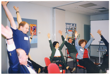 COL photo of a small group of people in a large room. One man is standing, while the others are sitting around some tables. They are all stretching their arms above and behind their heads, with their palms open and fingers spread out. In the background, there are two display boards on the on the walls with posters on them. One of the display boards also has some cut-out pieces of paper in the shape of feet and several photos pinned to the board. To the right of the frame, there is a freestanding slide projector screen on a stand with an overhead slide projector, also on a stand, in front of it.