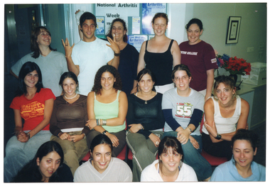 COL medium close up photo of a group of people sitting or standing across three rows. The people in the back row are standing; those in the middle row are sitting on chairs; and those in the front row are sitting on the floor. In the background, there is a display board with some posters on it. One of the posters contains the text, "National Arthritis Week".