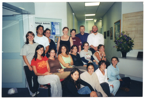 COL wide frame photo of a group of people sitting or standing across three rows. The people in the back row are standing; those in the middle row are sitting on chairs; and those in the front row are sitting on the floor. Behind them, there is a glass-fronted display board with some posters inside. One of the posters contains the text, "National Arthritis Week". On the opposite wall, there is a large floral bouquet on a hall table against the wall, and a large information sign hanging on the wall above it. There is a corridor behind them, with several framed pictures on the walls. The photo has been taken at a 45 degree angle.