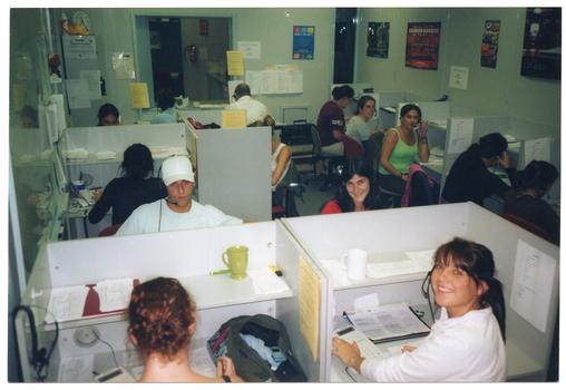 COL photo of a group of people sitting at small individual cubicles in an open office. Each person is wearing a telephone receiver headset except for one woman in the foreground, who has her telephone headset hanging on the short dividing wall of her cubicle. There is a mug on the shelves of the two cublicles in the foreground. There is a large whiteboard to the left of the frame, and several posters and printed pieces of paper on the walls around the room. In the background, an open double doorway leads to another room. This photo has been taken from a slightly higher vantage, looking down toward the subjects.