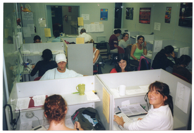 COL photo of a group of people sitting at small individual cubicles in an open office. Each person is wearing a telephone receiver headset except for one woman in the foreground, who has her telephone headset hanging on the short dividing wall of her cubicle. There is a mug on the shelves of the two cublicles in the foreground. There is a large whiteboard to the left of the frame, and several posters and printed pieces of paper on the walls around the room. In the background, an open double doorway leads to another room. This photo has been taken from a slightly higher vantage, looking down toward the subjects.
