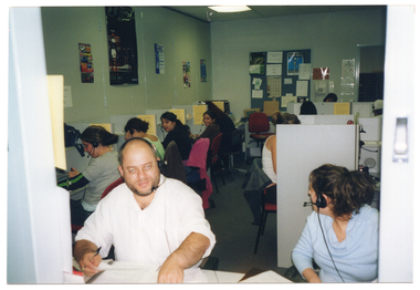 COL photo of a group of people sitting at small individual cubicles in an open office. Each person is wearing a telephone receiver headset. This photo was taken through the servery window from an adjoining room. There are posters on the walls, and a display board with posters and other information on it.