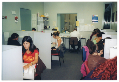 COL photo of a group of people sitting at small individual cubicles in an open office. One person is sitting at a small table against the wall beneath a servery window. Each person is wearing a telephone receiver headset. There is a wall shelf in the corner, and a few small posters or pictures and information sheets on the walls.