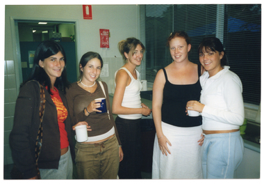 COL photo of five young women standing next to one another in a large kitchen. Three of the women are holding a mug in their hands. Behind them, there is a large window with thin venetian blinds above the kitchen sink.