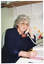 COL medium close up photo of a woman sitting at a desk, looking directly at the camera. She is wearing a navy blue pin-stripe jacket over a black top, with a yellow and pink flower appliqued at the top. She is holding a telephone receiver to her ear with one hand and, in her other hand, has a pen poised over a notebook. Behind her, there is a wooden shelf attached to the wall with brackets.