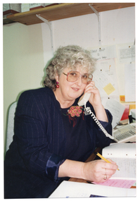 COL medium close up photo of a woman sitting at a desk, looking directly at the camera. She is wearing a navy blue pin-stripe jacket over a black top, with a yellow and pink flower appliqued at the top. She is holding a telephone receiver to her ear with one hand and, in her other hand, has a pen poised over a notebook. Behind her, there is a wooden shelf attached to the wall with brackets.