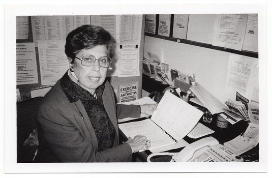 B&W photo of a woman sitting at a desk with a telephone, a desk calender, and miscellaneous stationery items. She is holding open an information booklet while facing the camera. There are typed pieces of paper on the wall next to and facing her, and various leaflets, brochures and other paperwork in small pigeonhole shelving on the wall. There is also a large noticeboard on the wall, above the pigeonholes.