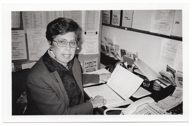B&W photo of a woman sitting at a desk with a telephone, a desk calender, and miscellaneous stationery items. She is holding open an information booklet while facing the camera. There are typed pieces of paper on the wall next to and facing her, and various leaflets, brochures and other paperwork in small pigeonhole shelving on the wall. There is also a large noticeboard on the wall, above the pigeonholes.