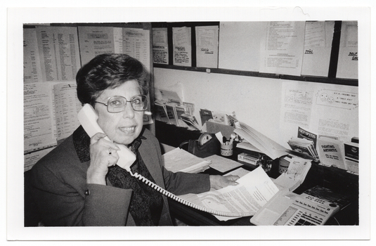 B&W photo of a woman sitting at a desk with a telephone, a desk calender, and miscellaneous stationery items. She is holding open an information booklet while facing the camera and holding the telephone receiver to her ear. There are typed pieces of paper on the wall next to and facing her, and various leaflets, brochures and other paperwork in small pigeonhole shelving on the wall. There is also a large noticeboard on the wall, above the pigeonholes.