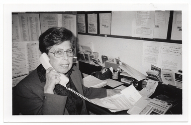 B&W photo of a woman sitting at a desk with a telephone, a desk calender, and miscellaneous stationery items. She is holding open an information booklet while facing the camera and holding the telephone receiver to her ear. There are typed pieces of paper on the wall next to and facing her, and various leaflets, brochures and other paperwork in small pigeonhole shelving on the wall. There is also a large noticeboard on the wall, above the pigeonholes.