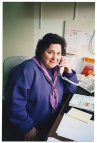 COL photo of a woman sitting at a desk, holding a telephone receiver to her ear. She is wearing a purple sweatshirt, with a purple scarf with a printed design. On the desk, there is an exercise book and two notepads, as well as a book titled 'Rheumatism'. On the wall beside her, there is some printed paper pinned to a noticeboard.
