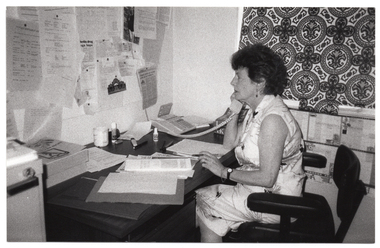 B&W medium close up photo of a woman sitting at a desk. She is holding a telephone receiver to one ear. On the desk, there are various papers and folders, two Yellow Pages telephone directories, as well as other office-related paraphanalia such as a glue stick, correction fluid, and a highlighter felt pen. The desk is against the wall, in the corner. There is a large noticeboard on the wall, with typed and printed papers pinned to the board.