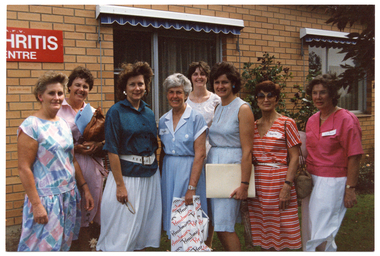 COL photo of a group of women standing outside a brick building. On the exterior wall of the building, there is a red sign with white text, "A.F.V. ARTHRITIS CENTRE" (partially obscured). There are two large rectangular windows, above which there are some pelmets, each with a blue and white striped retractable awning underneath. There is a large rose bush against the exterior brick wall between the two windows.