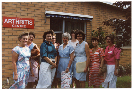 COL photo of a group of women standing outside a brick building. On the exterior wall of the building, there is a red sign with white text, "A.F.V. ARTHRITIS CENTRE" (partially obscured). There are two large rectangular windows, above which there are some pelmets, each with a blue and white striped retractable awning underneath. There is a large rose bush against the exterior brick wall between the two windows.