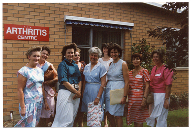 COL photo of a group of women standing outside a brick building. On the exterior wall of the building, there is a red sign with white text, "A.F.V. ARTHRITIS CENTRE" (partially obscured). There are two large rectangular windows, above which there are some pelmets, each with a blue and white striped retractable awning underneath. There is a large rose bush against the exterior brick wall between the two windows.
