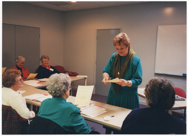 COL photo of a woman standing in a room in front of a group of women who are seated at tables. She is holding a piece of paper in her hand. Some of the women seated are also holding a piece of paper in their hands. The tables (partially obscured) have been arranged in an L-shape. Behind the woman standing, there is another smaller table with a telephone on top. In the background, there is a large whiteboard affixed to the wall.