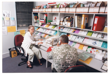 COL photo of two people sitting at two desks along a wall. The woman (facing the camera) is wearing a telephone receiver headset and the man (with his back to the camera) is using a telephone receiver handset. Above the desks, there are several shelves with brochures, folders, and other printed materials (booklets, etc) against the wall. Next to the window on the other wall, there are some brochures in paper pockets taped to the wall.