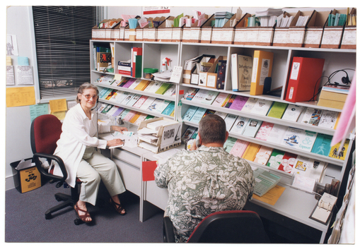 COL photo of two people sitting at two desks along a wall. The woman (looking directly at the camera) is wearing a telephone receiver headset and the man (with his back to the camera) is using a telephone receiver handset. Above the desks, there are several shelves with brochures, folders, and other printed materials (booklets, etc) against the wall. Next to the window on the other wall, there are some brochures in paper pockets taped to the wall.