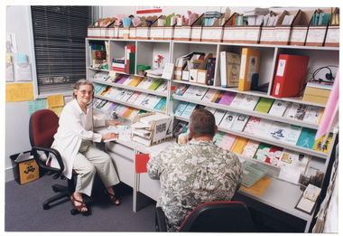 COL photo of two people sitting at two desks along a wall. The woman (looking directly at the camera) is wearing a telephone receiver headset and the man (with his back to the camera) is using a telephone receiver handset. Above the desks, there are several shelves with brochures, folders, and other printed materials (booklets, etc) against the wall. Next to the window on the other wall, there are some brochures in paper pockets taped to the wall.