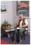 COL photo of a woman sitting on a chair next to a small square side table. On the table, there is a telephone, a potted plant, and a small book. The woman is holding the telephone receiver handset to her ear, with a pen in the same hand. She has a large notebook placed on her lap. There is a print or painting on the wall. Behind her, there is a window, through which a parked car and the house on the adjoining property are evident.