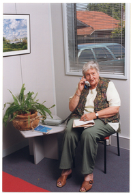 COL photo of a woman sitting on a chair next to a small square side table. On the table, there is a telephone, a potted plant, and a small book. The woman is holding the telephone receiver handset to her ear, with a pen in the same hand. She has a large notebook placed on her lap. There is a print or painting on the wall. Behind her, there is a window, through which a parked car and the house on the adjoining property are evident.