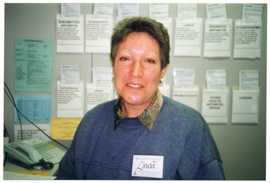 COL medium close-up photo of a woman sitting at the desk in an office. There is a multiple-line telephone system with a handset receiver on the desk. On the wall behind her, there are information sheets and various brochures tucked into paper pockets taped to the wall.