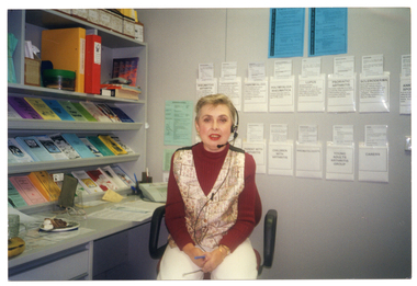 COL medium close up photo of a woman sitting at a desk in an office. She is wearing a telephone receiver headset. On top of the desk, there are bookshelves against the wall, containing brochures and folders. On the wall behind her, there are various brochures tucked into some paper pockets taped to the wall, with some information posters above. On the desk, there are also a small plate of cakes, a pair of spectacles, a banana and glass mug (partially obscured).