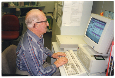COL photo of a man sitting at a computer desk with his hands poised over the keyboard. The computer monitor, which sits on top of a computer hard drive, displays a colour screen. There is a floppy disk placed on the hard drive. Behind him, there is a tall metal cabinet, an office chair, and a desk against the wall, with some shelving above it.