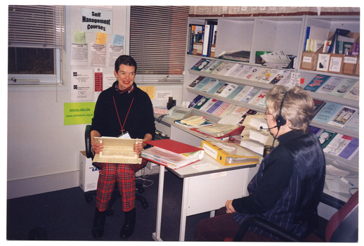 COL photo of two women seated at two desks against the wall. One woman (facing the camera) is holding a wooden book rest on her lap. The other woman (with her back to the camera) is wearing a telephone receiver headset. Above the desks, there are several shelves with brochures, folders, and other printed materials. There are two windows with thin venetian blinds on the other wall. On the wall between the windows, there is a laminated sign that says, "Self Management Courses". Below the sign, there are some brochures in paper pockets taped to the wall, as well as some other signs and information sheets.