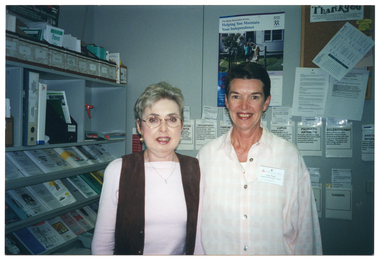 COL medium close up photo of two women standing close together in a room. Behind them, there are shelves containing brochures, folders, and other miscellaneous items on one wall, and on the other wall, brochures tucked into paper pockets taped to the wall. There is also a poster and a cork display board on the wall.