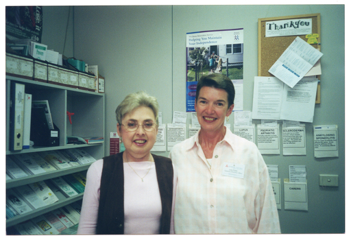 COL medium close up photo of two women standing close together in a room. Behind them, there are shelves containing brochures, folders, and other miscellaneous items on one wall, and on the other wall, brochures tucked into paper pockets taped to the wall. There is also a poster and a cork display board on the wall.