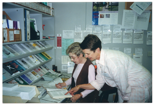 COL photo of two women in an office. One woman is sitting down at a desk with an open book, while the other woman is standing behind her, leaning over her shoulder and pointing to something in the book. On top of the desk, there is a telephone, a desk calendar, a thick book, and a printed paper form. On top of the desk, against the wall, there are bookshelves containing brochures and folders. On the other wall, there are brochures tucked into paper pockets taped onto the wall, a poster, and a cork noticeboard with some information sheets pinned onto it.