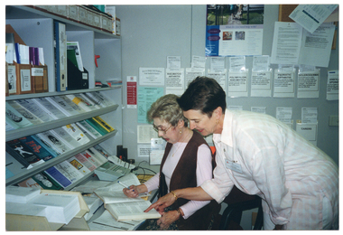 COL photo of two women in an office. One woman is sitting down at a desk with an open book, while the other woman is standing behind her, leaning over her shoulder and pointing to something in the book. On top of the desk, there is a telephone, a desk calendar, a thick book, and a printed paper form. On top of the desk, against the wall, there are bookshelves containing brochures and folders. On the other wall, there are brochures tucked into paper pockets taped onto the wall, a poster, and a cork noticeboard with some information sheets pinned onto it.
