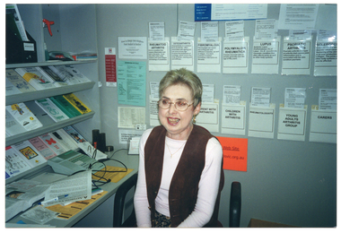 COL photo of a woman sitting at a desk in an office. On top of the desk, there are are shelves of brochures and folders against the wall. Behind her, there are brochures tucked into paper pockets taped onto the wall.