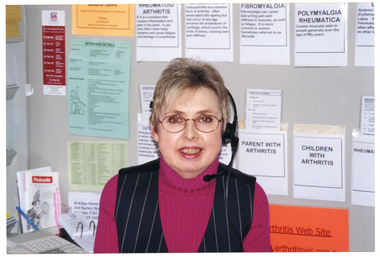 COL medium close up photo of a woman sititng at a desk. She is wearing a telephone headset. On the wall behind her, there are some information sheets on the wall, and various brochures tucked into paper pockets taped to the wall.