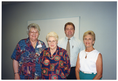 COL medium close up photo of four people standing close together in a room. There is a large whiteboard affixed to the wall behind them.
