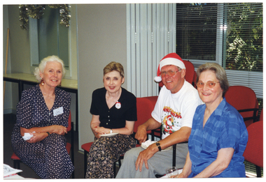 COL medium close up photo of four people sitting close together on some chairs in a room. Behind them, there is a set of bi-fold doors folded back into the open position, and a few stacks of chairs in front of a large window with venetian blinds. Beyond the open bi-fold doors, in an adjoining room, there are two rectangular tables pushed up against the wall beneath a servery window. A garland of thick tinsel is handing above the servery window. There is a viw of some trees evident through the window.