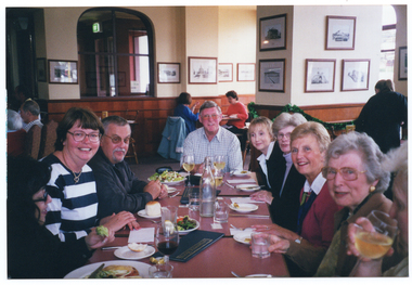 COL photo of a group of people sitting at a rectangular wooden table at in a large dining room. On the table, there are some meals on large plates, bread rolls on small plates, some glasses of wine or water, some water bottles, and some menus. In the background, there are other people seated at tables, dining. There are several framed pencil drawings displayed on the walls.