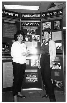 B&W photo of two women standing in front of a large multipanel display board titled, "Arthritis Foundation of Victoria". The board contains text and Photographs about the Foundation and its services.