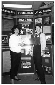 B&W photo of two women standing in front of a large multipanel display board titled, "Arthritis Foundation of Victoria". The board contains text and Photographs about the Foundation and its services.