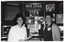 B&W medium close-up photo of two women standing in front of a large multipanel display board titled, "Arthritis Foundation of Victoria". They are both smiling. The display board contains text and Photographs about the Foundation and its services. The Photograph image is blurry.