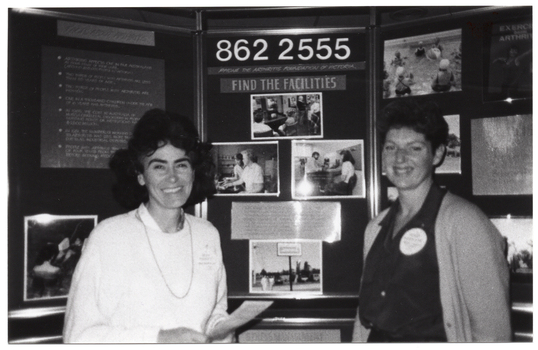 B&W medium close-up photo of two women standing in front of a large multipanel display board titled, "Arthritis Foundation of Victoria". They are both smiling. The display board contains text and Photographs about the Foundation and its services. The Photograph image is blurry.