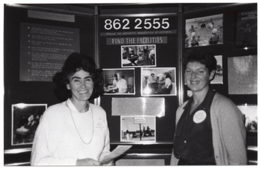 B&W medium close-up photo of two women standing in front of a large multipanel display board titled, "Arthritis Foundation of Victoria". They are both smiling. The display board contains text and Photographs about the Foundation and its services. The Photograph image is blurry.