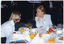 COL photo of two women sitting at a round table covered in a white tablecloth. On the table, there are plates of sandwiches and other food, wine glasses (some filled with orange juice and some empty), a carafe of orange juice, a jug of water, cups and saucers, and milk jugs. Also on the table, in the foreground, there is a framed photo or certificate (partially obscured). In the background, to the left of the frame, a waiter holding tea and coffee pots is approaching. In the background, to the right of the frame, a musician (partially obscured) is playing an electric keyboard.