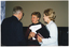COL photo of three people standing in a room, chatting. In the background, to the left of the frame, there is a white vertical "Move it or Lose it" banner (partially obscured). The lower half of the wall is covered in dark wood panelling.
