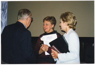 COL photo of three people standing in a room, chatting. In the background, to the left of the frame, there is a white vertical "Move it or Lose it" banner (partially obscured). The lower half of the wall is covered in dark wood panelling.