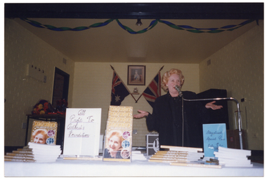COL photo of a woman speaking into a microphone on a stand. She is standing behind a long rectangular table. On the table are stacks and displays of copies of a book titled "Stagestruck". Between two book displays is a handwritten sign on white paper with the text "All profits to Arthritis Foundation". On the other side of one of the displays is another handwritten sign on blue paper, with the text  "Stagestruck Special Price Only $..." (partially obscured). In the background, there is a framed print of Queen Elizabeth II (1926-2022) on the wall, with a small silver urn on a shelf and two flags below it. There are two strings of blue and green streamers across the ceiling.